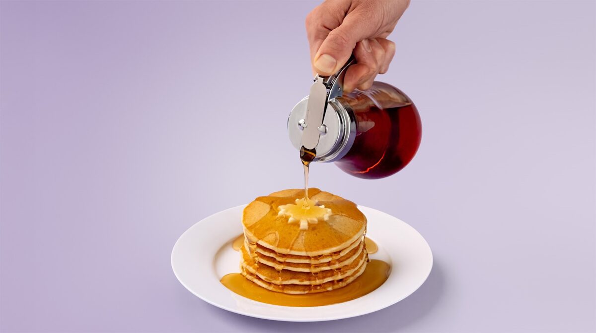 Syrup pouring over a stack of pancakes with a pat of butter in the shape of a Canadian maple leaf