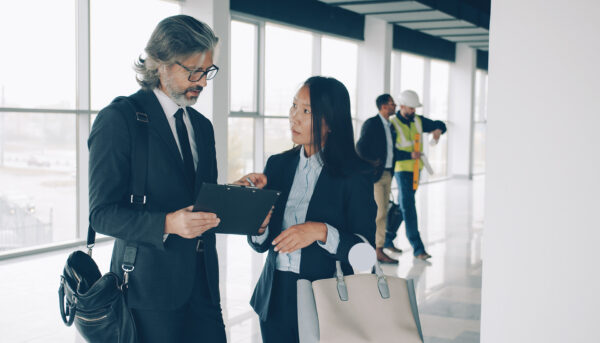 Multi-ethnic business partners man and woman are talking in modern building while workers in uniform are busy in background.