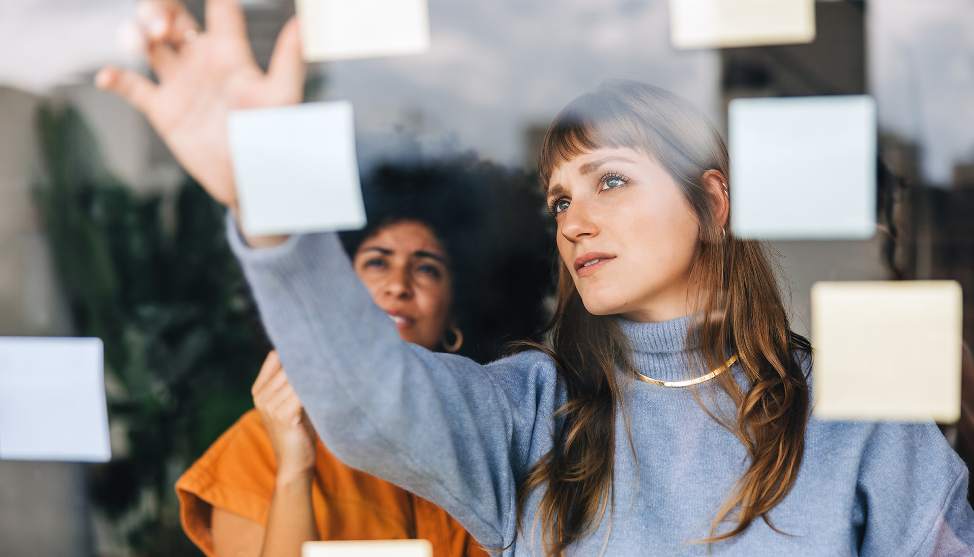 two young women standing in front of a clear bulletin board brainstorming ideas with sticky notes