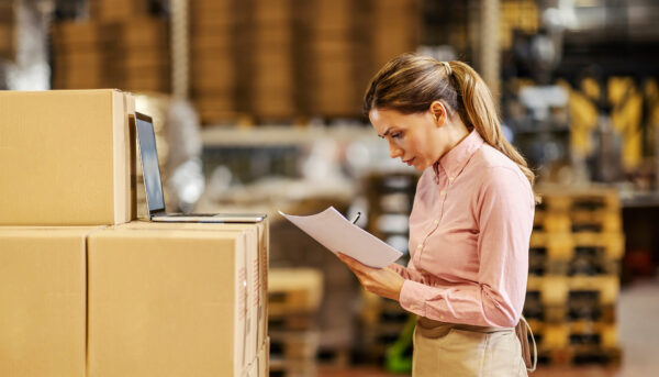 woman in a pink shirt verifying documents in a warehouse. She is standing on front of a stack of boxes with a laptop on top.