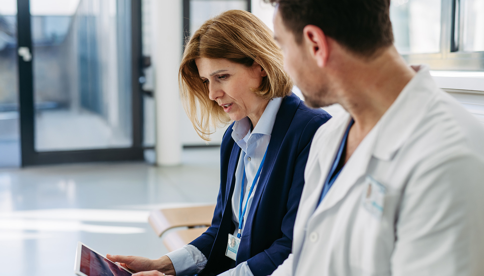 female pharmaceutical representative speaking to male doctor in a white coat and working on tablet.