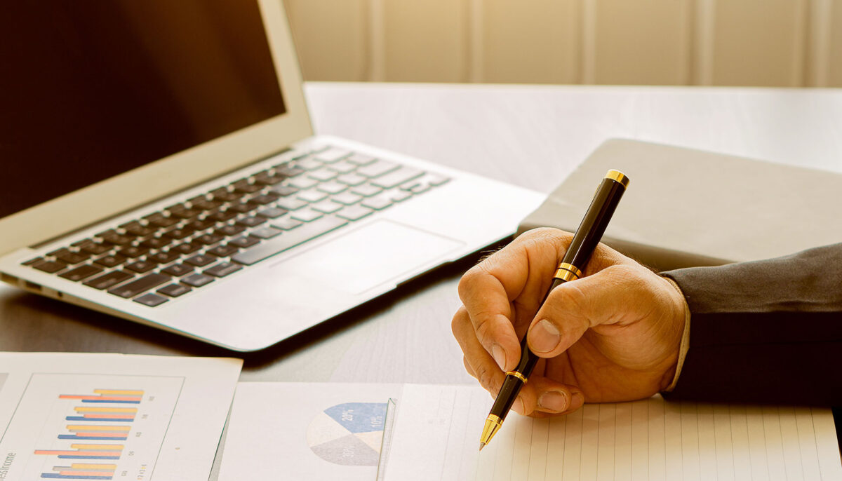 hand writing on notepad with charts and open laptop in the background