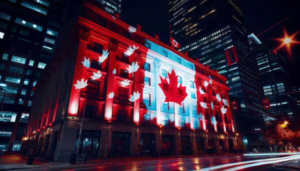Historic building illuminated in red and white with large Canadian maple leaf projections across the facade at night, surrounded by downtown high-rise buildings. Canadian flags hang from the exterior, and light trails from passing traffic are visible along the street.