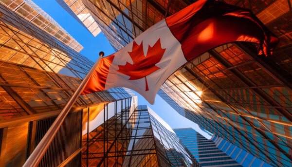 Large Canadian flag waving on a flagpole between modern glass office towers, photographed from a low angle looking upward.