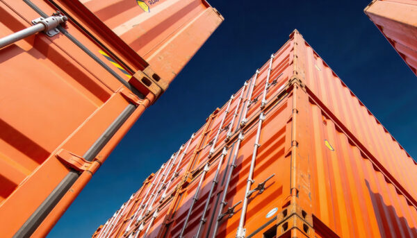 ow-angle view of bright orange shipping containers stacked in a storage yard, with the containers towering overhead against a clear blue sky.