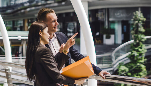 Two professionals stand on a modern indoor walkway overlooking a retail atrium. One person points into the distance while the other holds a large orange folder.