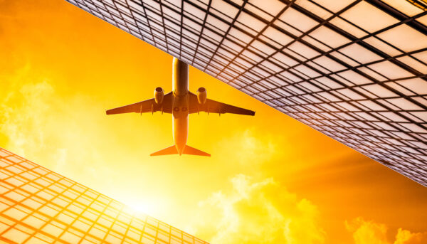 Low-angle view of an airplane flying between two modern glass office towers against a bright orange and yellow sky.