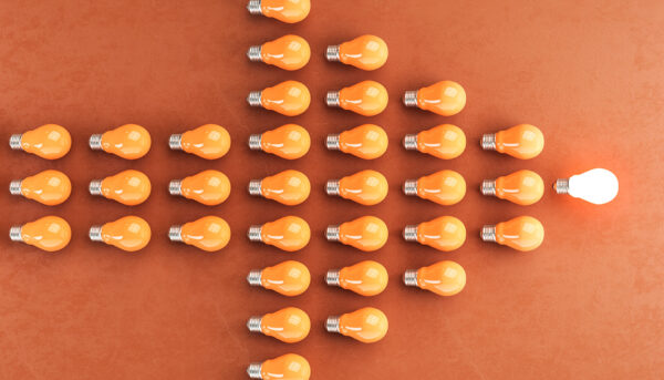 Overhead view of multiple orange light bulbs arranged in the shape of a right-pointing arrow against an orange background. One illuminated white light bulb stands at the tip of the arrow.