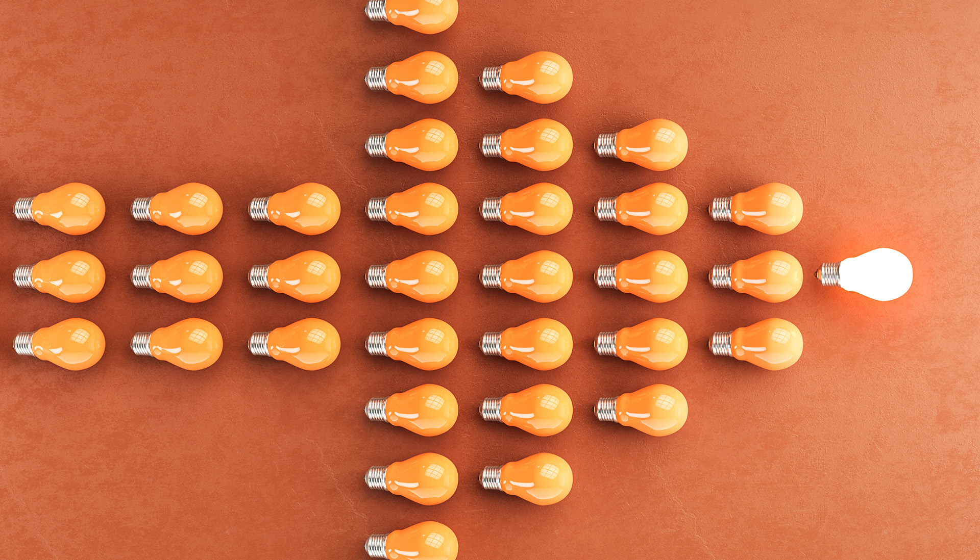 Overhead view of multiple orange light bulbs arranged in the shape of a right-pointing arrow against an orange background. One illuminated white light bulb stands at the tip of the arrow.