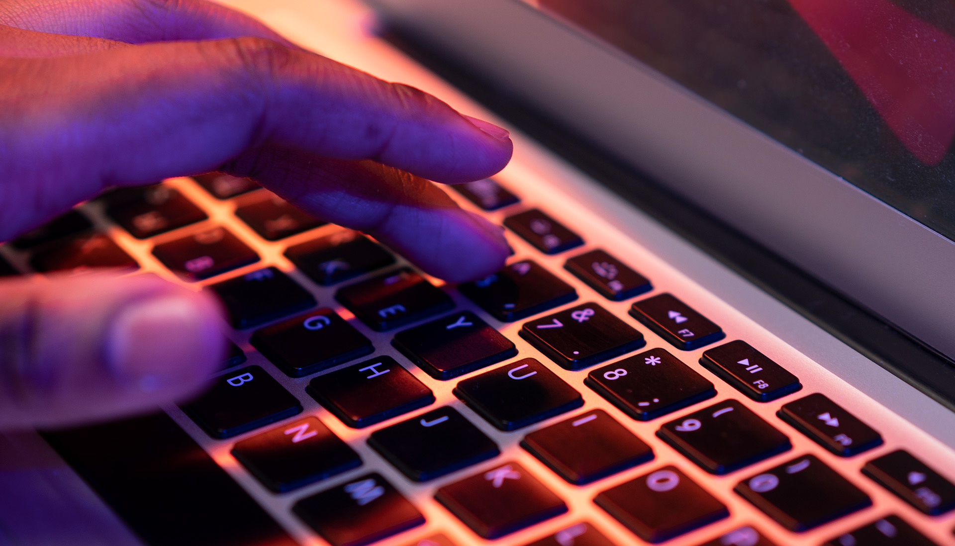 Close-up of a hand typing on a laptop keyboard illuminated by vivid purple and orange lighting.