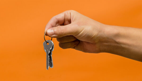Close-up of a hand holding a key ring with two silver keys against a bright orange background.