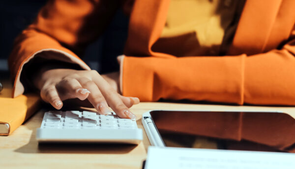 Close-up of a person’s hand pressing buttons on a calculator at a desk. A tablet, documents and a notebook are nearby