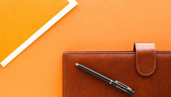 Brown leather planner with a pen resting on top, positioned on a bright orange surface.