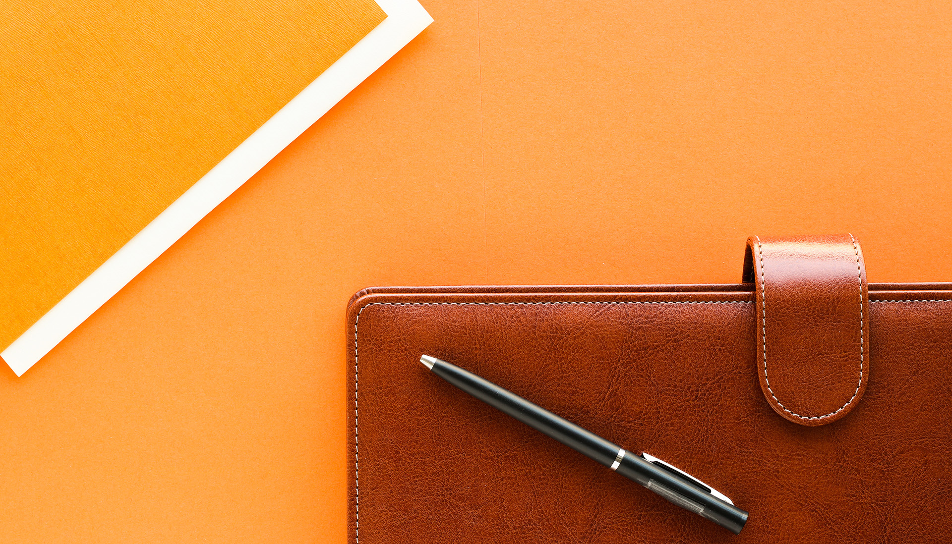Brown leather planner with a pen resting on top, positioned on a bright orange surface.