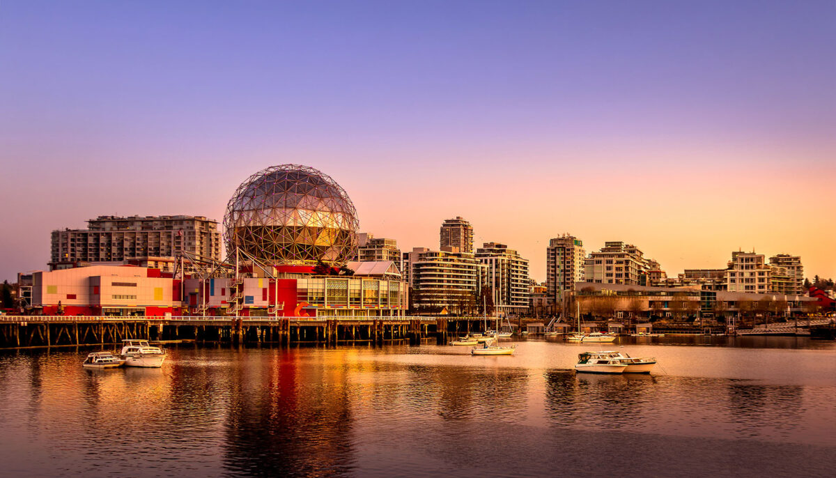Sunset view of False Creek in Vancouver, featuring Science World’s illuminated geodesic dome along the waterfront, surrounded by modern high-rise buildings. Several small boats rest on the calm water, reflecting the warm pink and orange hues of the evening sky.